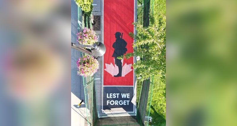 The red, black, and white design features a soldier and maple leaf, along with the words “Lest We Forget.”